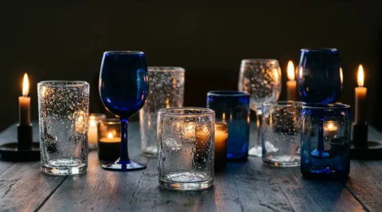 Verreries artisanales bullées transparentes et bleutées disposées sur une table sombre éclairées à la lueur de bougies