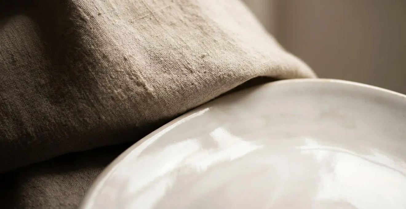 Contraste entre la texture mate d'un tissage de lin brut et la surface brillante d'une assiette en faïence émaillée