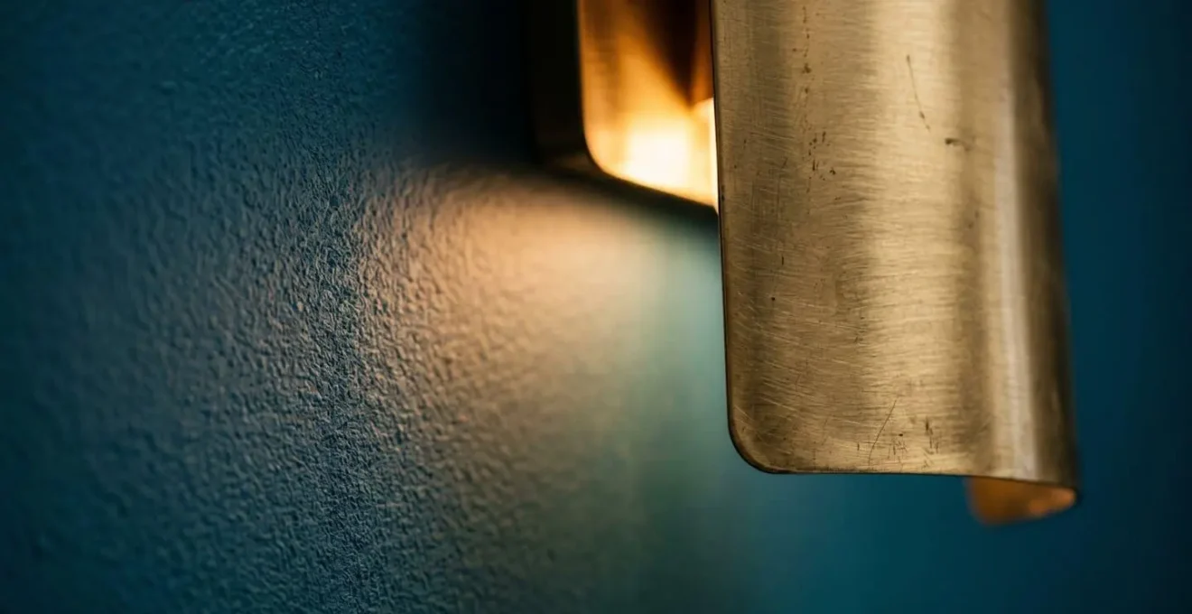 Détail d'une applique en laiton brossé projetant une lumière chaude sur un mur bleu pétrole mat